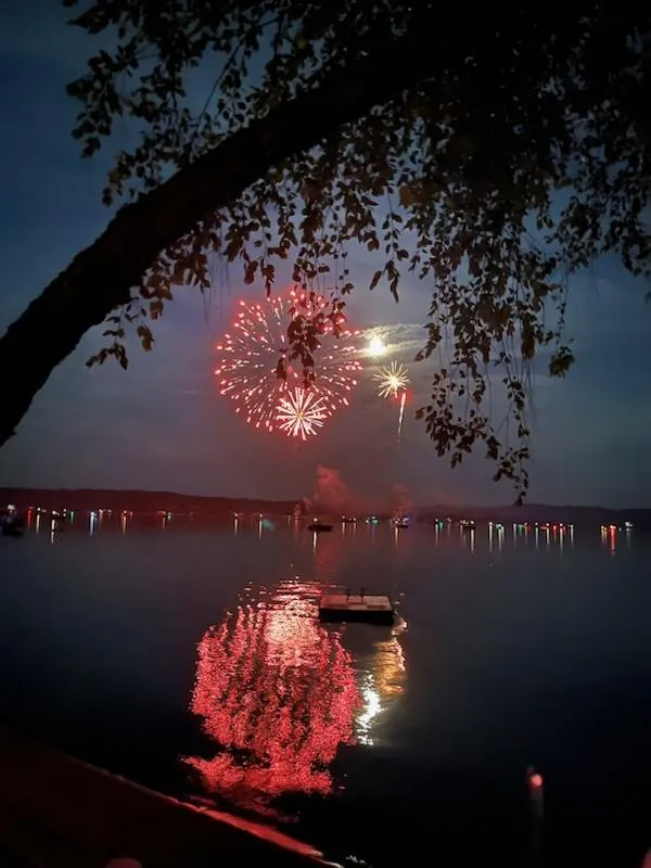 Red fireworks reflected in Lake Arbutus beneath shoreline trees.